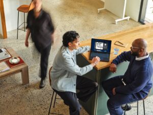 Two people in conversation while reviewing information on a laptop in a professional setting.