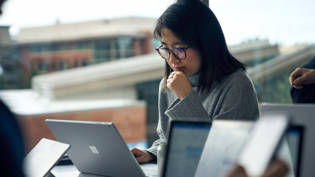 Woman with glasses sitting at a community table in common area reading on a Surface laptop.